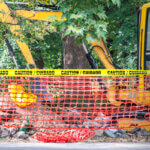 Excavator digging by a tree and concrete with barricade tape fencing in the site.