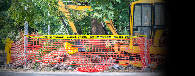 Excavator digging by a tree and concrete with barricade tape fencing in the site.